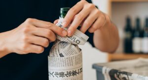 ワインセラーがない場合の代替保管法として、日本人の手によってワインボトルが丁寧に新聞紙で包まれている様子。